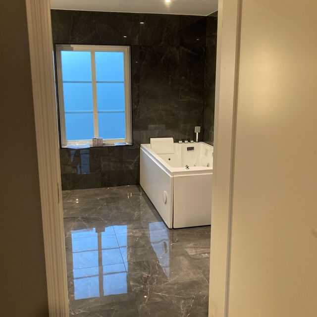 Badrum med blankt stengolv, vitt badkar, fönster med spröjs