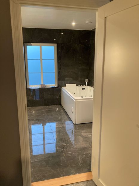 Badrum med blankt stengolv, vitt badkar, fönster med spröjs