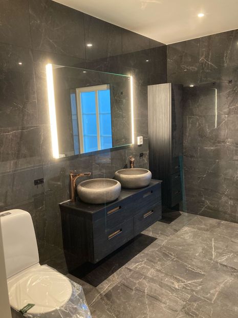 Grått badrum med blankt stengolv, svart kommd med två runda gråa stenhandfat, stor spegel med sidobelysning